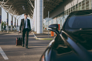 Full length young traveler businessman young man 20s wear black dinner suit going walk outside at international airport terminal with suitcase to car booking taxi. Air flight business trip concept