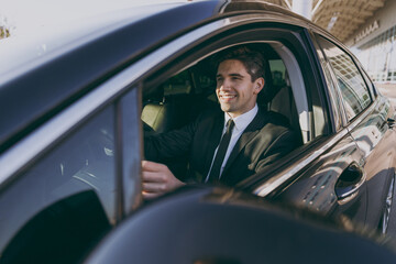 Side profile view smiling young driver smiling businessman man 20s wear black dinner suit driving car taxi hold steering wheel look camera Vehicle transport traffic lifestyle business trip concept.