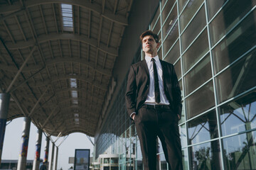 Bottom view confident young traveler brunet businessman man in black classic tie suit standing outside at international airport terminal look aside. People air flight business trip lifestyle concept.