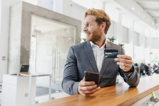Man businessman manager male client in classic suit standing at office table look aside use mobile cell phone credit bank card in dealership salon store office indoor Business lifestyle sales concept