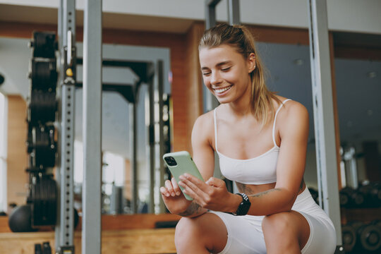 Young happy strong sporty athletic sportswoman woman 20s in white sportswear warm up training sitting near treadmill trainers using mobile cell phone in gym indoors. Workout sport motivation concept