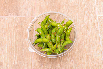 Edamame pods with salt flakes and hot sauce inside a transparent plastic bowl for home delivery