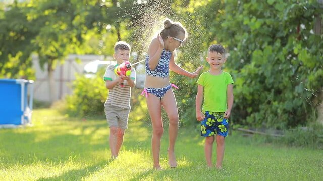 three fun kids boys and girl playing with water guns on green lawn in the backyard at hot summer day in slow motion. swimming pool on background