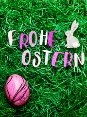 Vertical photo with letters saying "Happy Easter" in German ("Frohe Ostern"), a pink marbled egg and a decorative little bunny on green easter grass.