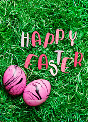 Vertical photo with letters saying "Happy Easter" and two pink-black marbled eggs on green easter grass with copy space to add text.