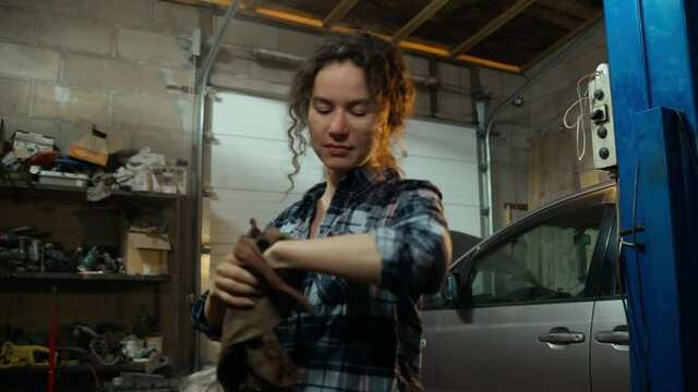 curly-haired millennial girl in a blue plaid shirt in a garage or at a service station wipes her hands of engine oil. Hand work. Car repair in the home garage. A woman at a man's work. 