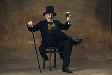 Elderly gray-haired man, gentleman, aristocrat or actor drinking beer isolated on dark vintage...