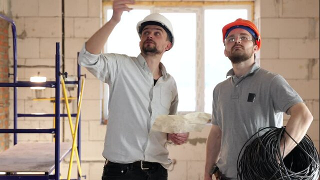 In the middle of the room being renovated, two construction workers in helmets are standing and discussing.