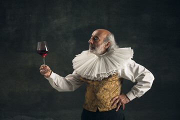 Elderly gray-haired man, medieval hystorical person, actor drinking wine isolated on dark vintage background. Retro style, comparison of eras concept. - obrazy, fototapety, plakaty