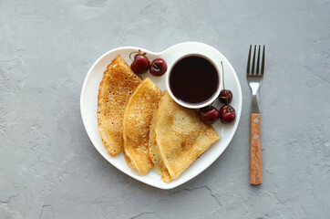 Pancakes with cherry jam and berries for breakfast in a white heart-shaped plate on a gray background. Top view. Concept: romantic breakfast.
