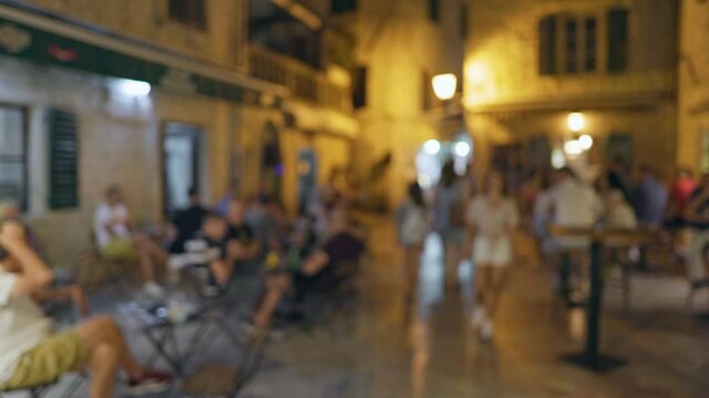 Defocused shot of people walk and sit in street cafes in an old European town. Unrecognizable people chat with a glass of wine and relax on the pedestrian street of the town in the evening. Cozy