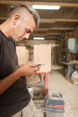 Carpenter artisan resting in workshop and scrolling on smartphone. Handsome handicraftsman texting message and tapping on phone