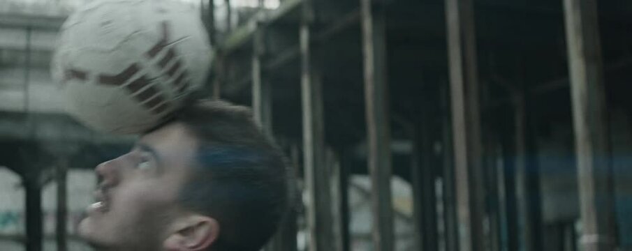 Anamorphic shot of young male athlete tossing up soccer ball and balancing it on his head in abandoned building