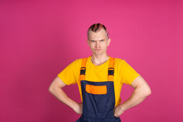 European handsome young man in yellow t shirt and overalls on pink background worker angry look to camera hands on waist