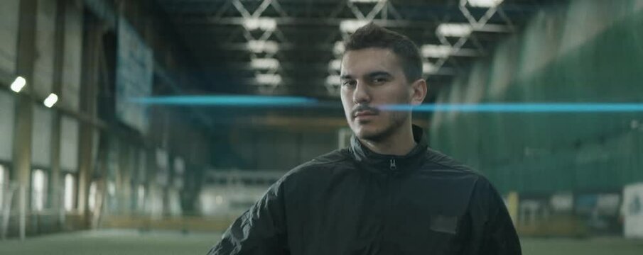 Zoom in anamorphic shot of handsome soccer player in sportswear holding ball looking at side and then posing for camera with confidence while standing on indoor football field