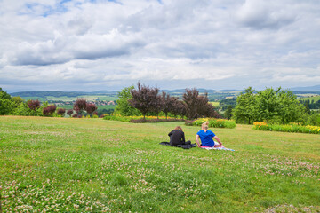 Piknik z przyjaciółmi na trawiastym wzgórzu z widokiem na cały świat, drzewa, kwiaty,, pochmurne niebo.