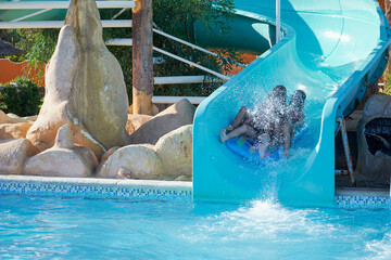 Dzieci na zjeżdżalni w hotelowym parku wodnym z dużą prędkością wpadają do basenu wśród rozprysków wody i piany. - obrazy, fototapety, plakaty