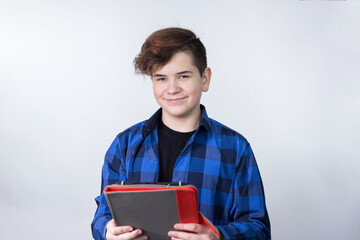 A teenage boy stands on a light background with a computer tablet in his hands. He is Caucasian. He's happy. The concept of education and online learning.