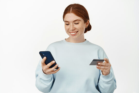 Image of smiling young redhead woman register her credit card in application, looking at mobile phone screen, holding discount card, standing against white background