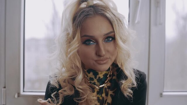 The charismatic young woman, blonde, with curls, shrill make-up, chicly raises her eyebrows, looks sexy and displays a small smile. Female energy. Beauty industry. Artistic concept