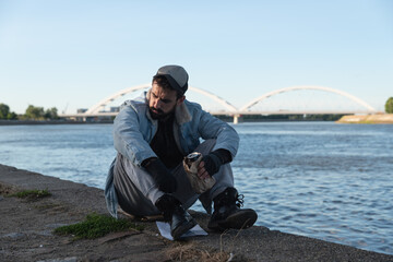 Young depressed suicidal drunk homeless man sitting under the bridge drinking alcohol drink and...