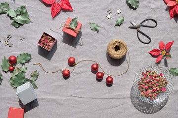 Christmas flat lay on natural beige linen textile. Gift boxes, red poinsettia, red baubles, winter decor. Dry rose tea leaves. Hemp cord, scissors, red and dark green poinsettia leaves .