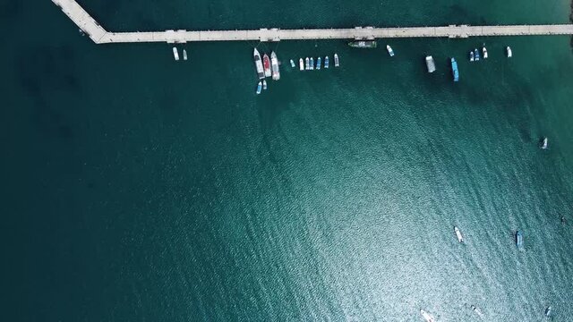Aerial shot. A beautiful up-down view of the pier in the blue ocean water. Fishing boats and yachts are floating next to it. Amazing reflection of the sunlight in the water.