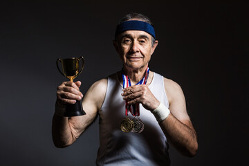 Elderly athlete wearing white sleeveless shirt, with sun marks on his arms, with three medals on his neck, with a trophy in his hands, on a dark background. Sports and victory concept.