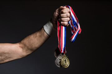 Fototapeta na wymiar Arm of elderly athlete holding three gold, silver and bronze medals on a dark background. Sports and victory concept.