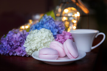 Obraz na płótnie Canvas Pink marshmallow on a white saucer with multicolored hydrangea flowers and a cup against the background of evening lights. Summer evening, holiday mood concept.