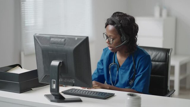 Black Woman Doctor Wear White Coat, Glasses And Headset Talking To Client Use Videocall. Due Coronavirus Pandemic Outbreak Therapist Working Remotely Provide Help To Clinic Patients By Video Call