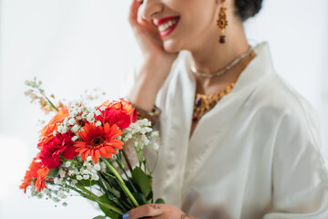 cropped view of cheerful indian bride holding bouquet of flowers on white