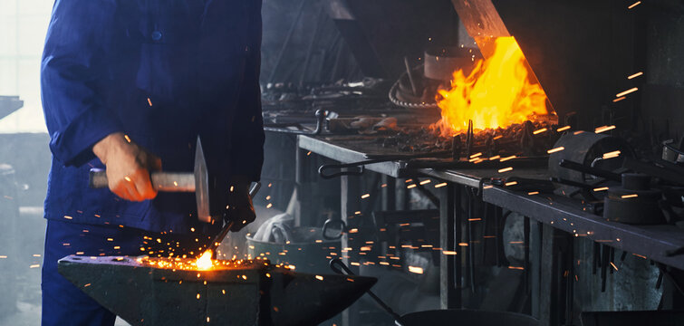 Close up of experienced blacksmith forging metal with heavy hammer on anvil. Man in blue overalls working with steel heated to high temperature.