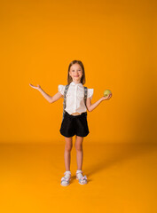 a little schoolgirl in a uniform and with a backpack stands and holds an apple on a yellow background with a copy of the space