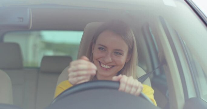 Happy young caucasian woman is driving a car on a road, smiling and showing okey. Car's interior. Front view. Close up.
