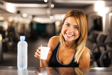 happy girl with bottle of water and a coffee cup resting in a generic out of focus fitness club.