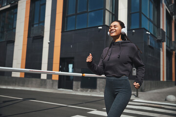 Smiling joyous sportswoman in wireless headphones and activewear jogging ahead