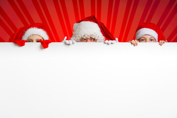 satna claus with funny children in red christmas gnome hats peeking out from behind a blank white...