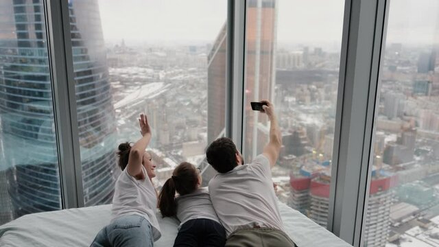 An authentic shot of a happy family taking a selfie or video call to their father or relatives in bed in a modern apartment overlooking the city. Technology concept, new generation