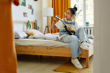 Full length portrait of trendy teenage girl playing ukulele while sitting on bed in cozy room