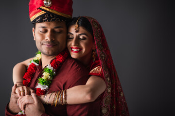cheerful indian bride in sari hugging bridegroom in turban isolated on grey