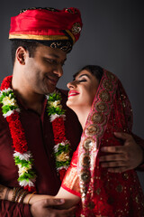 Indian man in turban hugging and looking at happy bride in traditional headscarf isolated on grey