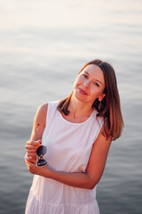 Portrait of a woman at sunset. A happy, sweet woman poses at sunset holding sunglasses near the sea on vacation on a trip. 