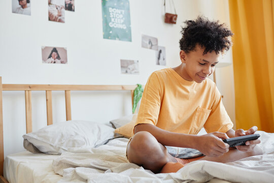Full length portrait of mixed-race teenage boy sitting on bed and playing mobile games via smartphone, copy space