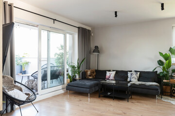 Interior of living room with wooden floor, big window and comfortable sofa with pillows. Room with balcony in modern apartment.