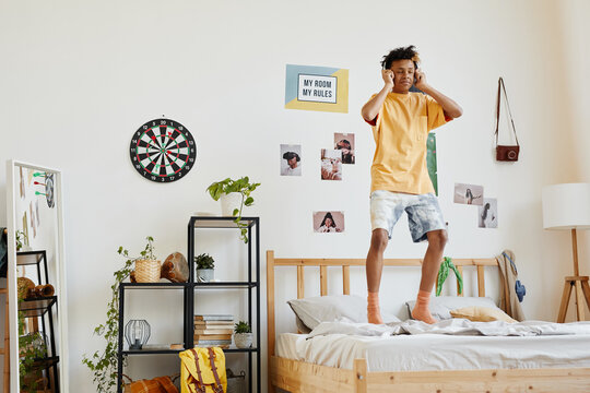 Wide angle portrait of mixed-race teenage boy jumping on bed while listening to music with eyes closed, copy space