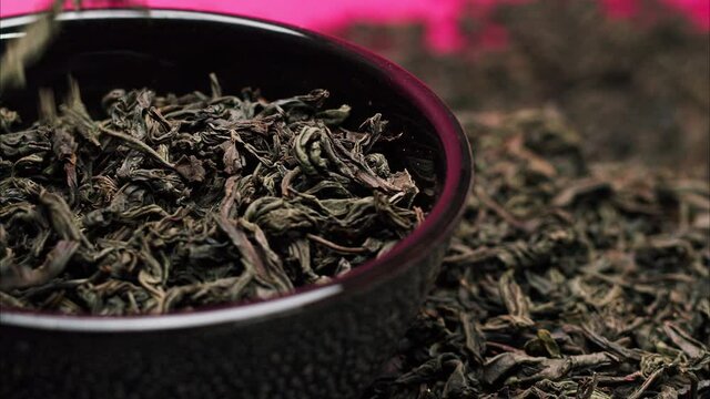 Close-up of falling down dried tea leaves into rotating black bowl on pink background. Shooting of ceylon tea in studio. Macro footage of pouring out brewed tea. Food and drink cooking video. 