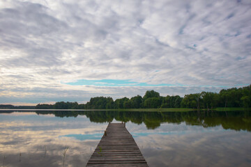 Lake with an old, long wooden footbridge in the center of the photo. Above the lake there are thick clouds, in the water you can see the reflection of clouds and trees