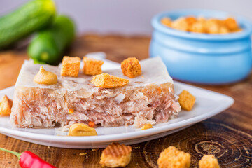 Beef and pork jellied meat on a white saucer. Cold appetizer with pieces of rusks and cucumber. Close-up.