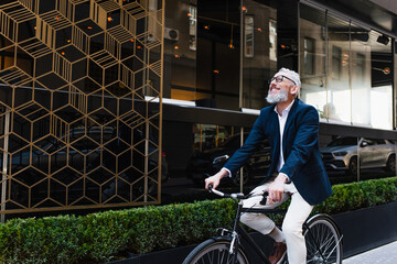 joyful middle aged man in blazer and glasses riding bicycle on modern urban street